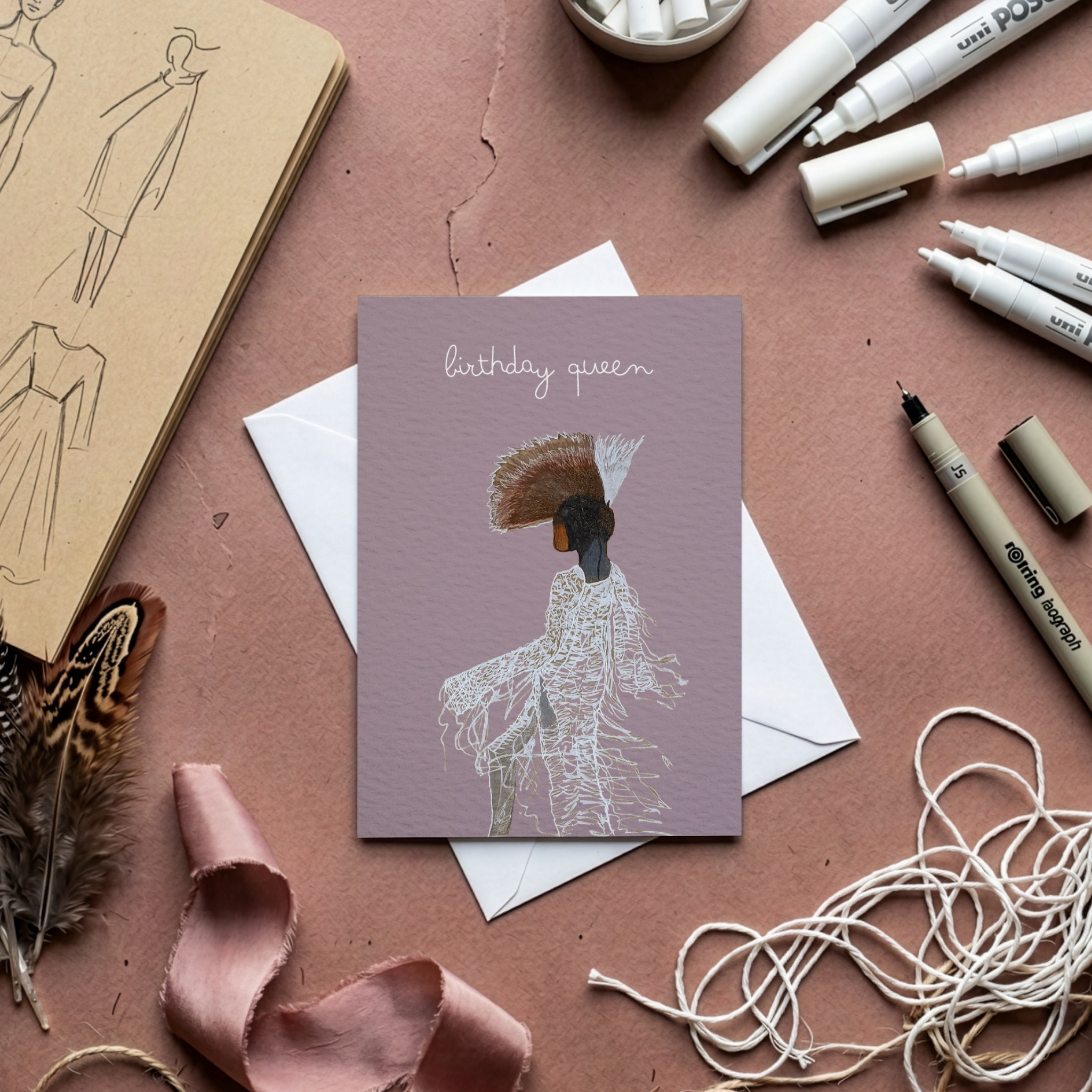 Greeting card with 'birthday queen' text and illustration on a pink surface with stationery items.