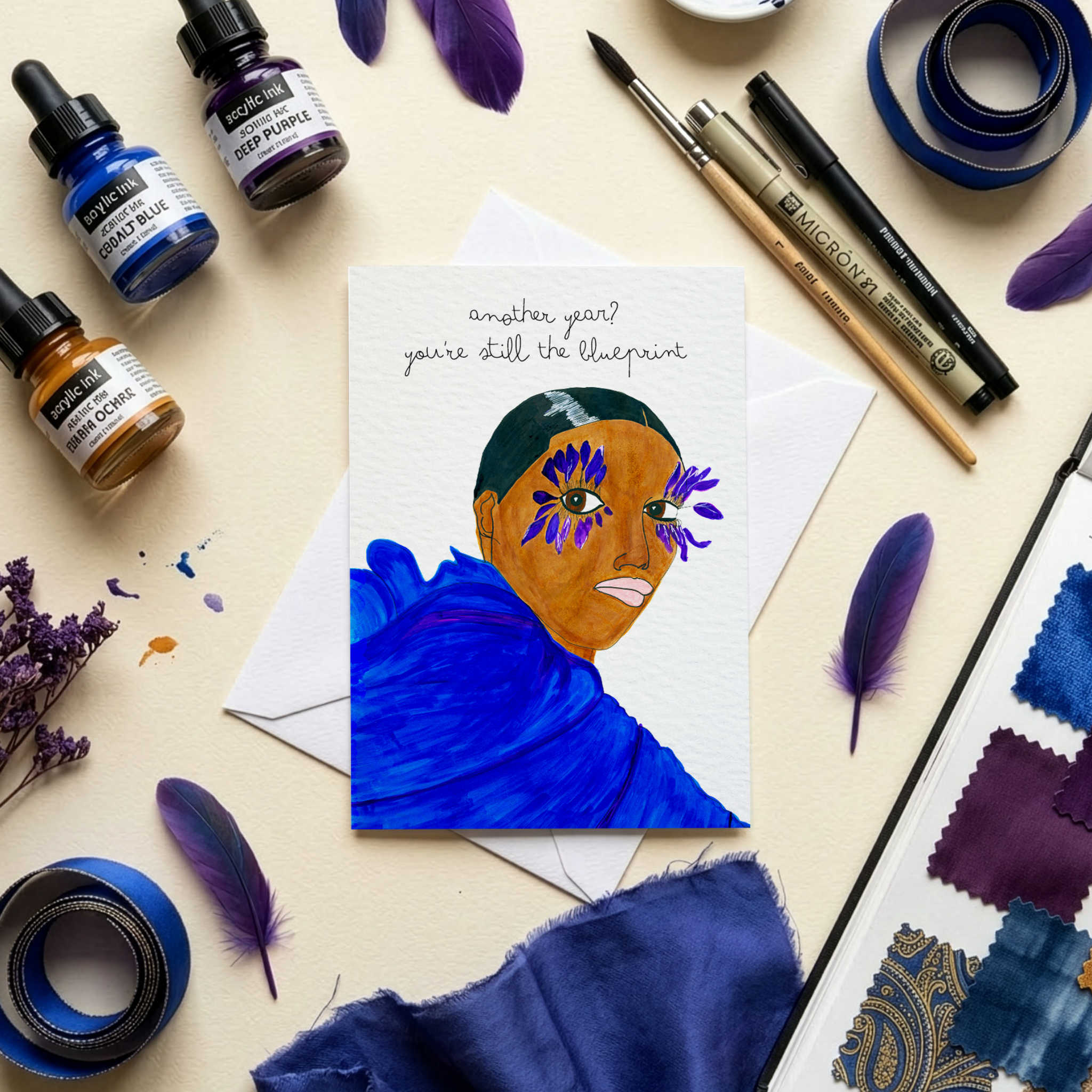 Colorful art supplies including paint bottles, feathers, and a fashion illustrated greeting card on a beige background.
