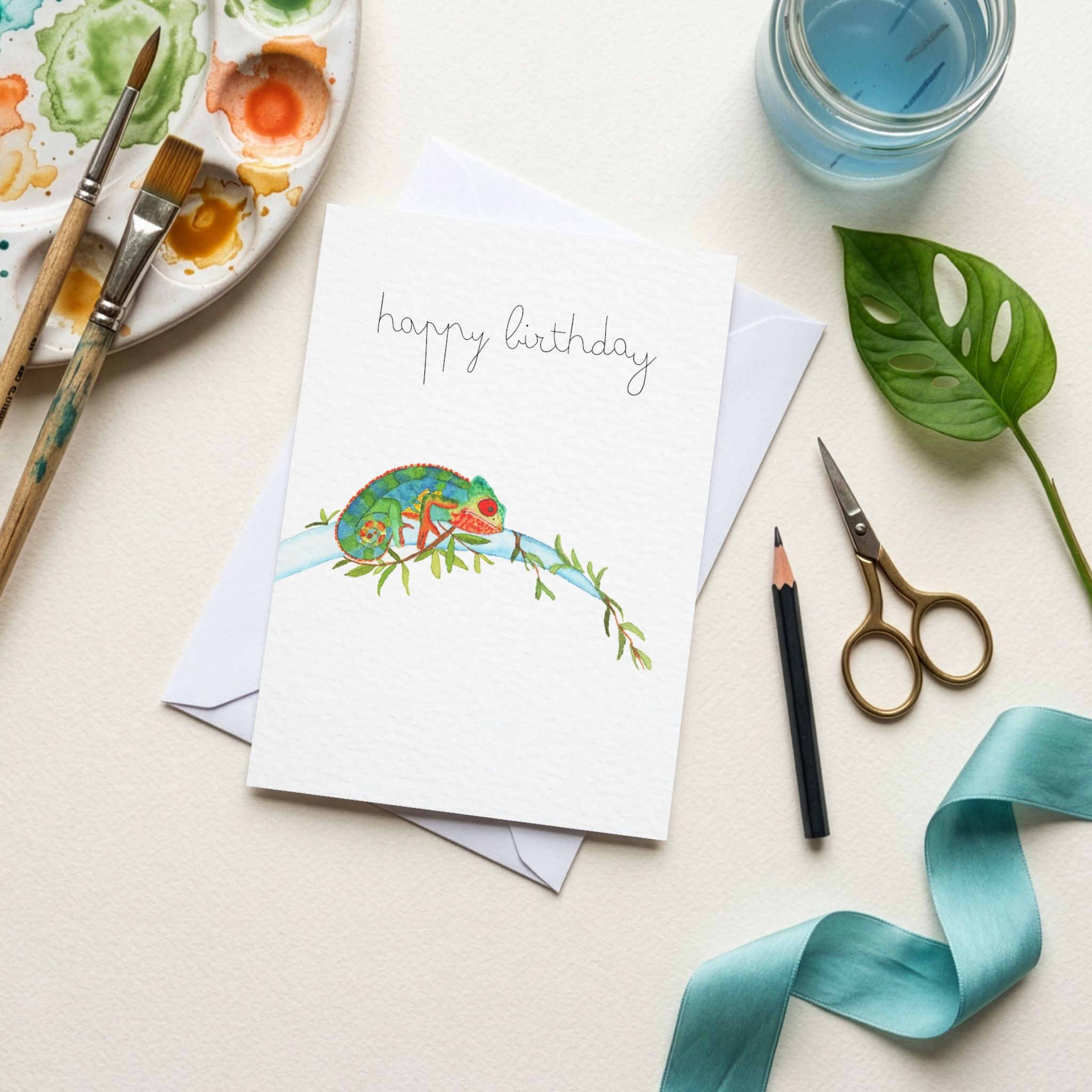 Happy birthday card with a chameleon watercolour illustration on a table with art supplies.
