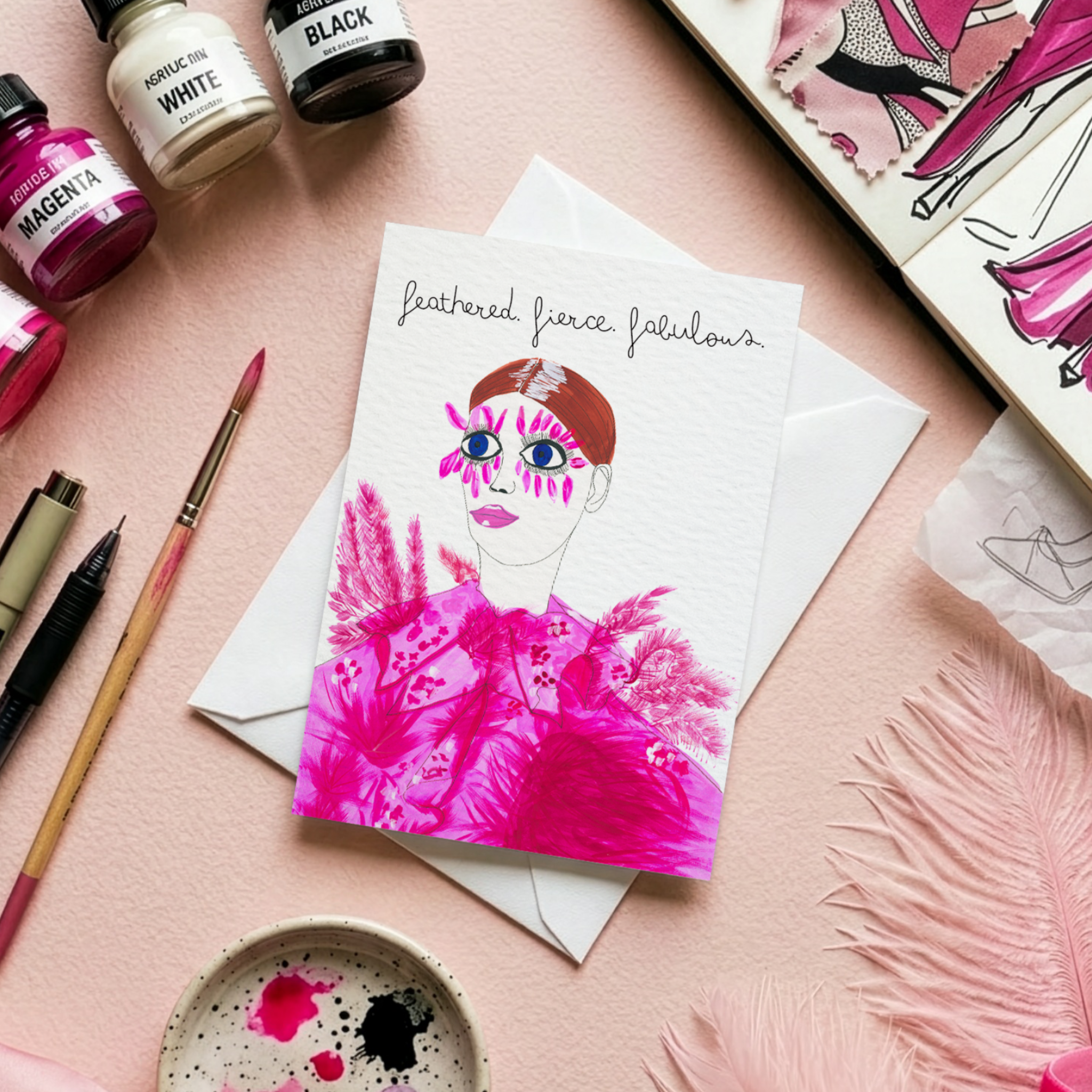 Art supplies including paint bottles, brushes, and a fashion illustration greeting card of a woman in a pink dress on a pink surface.
