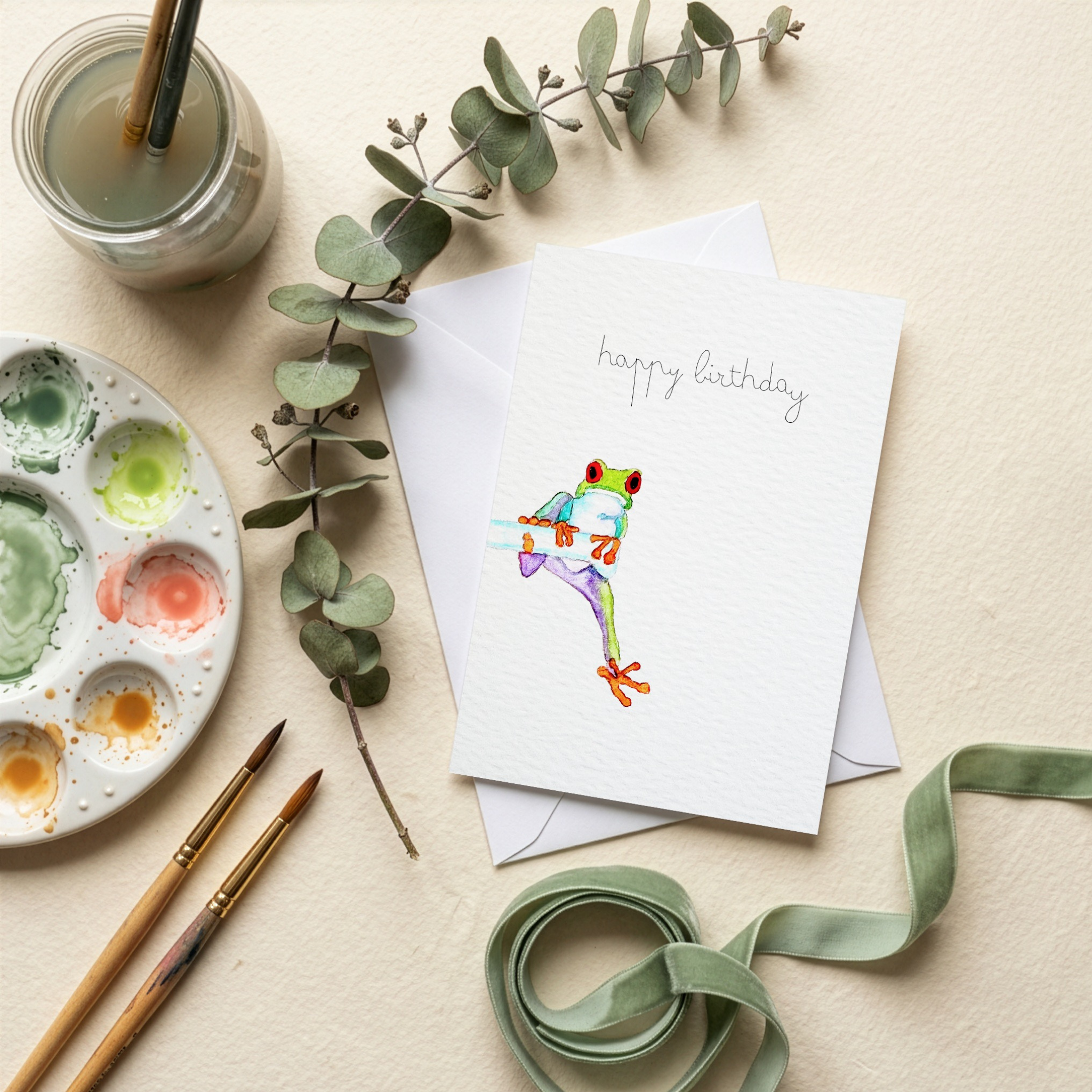 Greeting card with a colorful frog watercolour illustration on a table with art supplies and eucalyptus leaves.