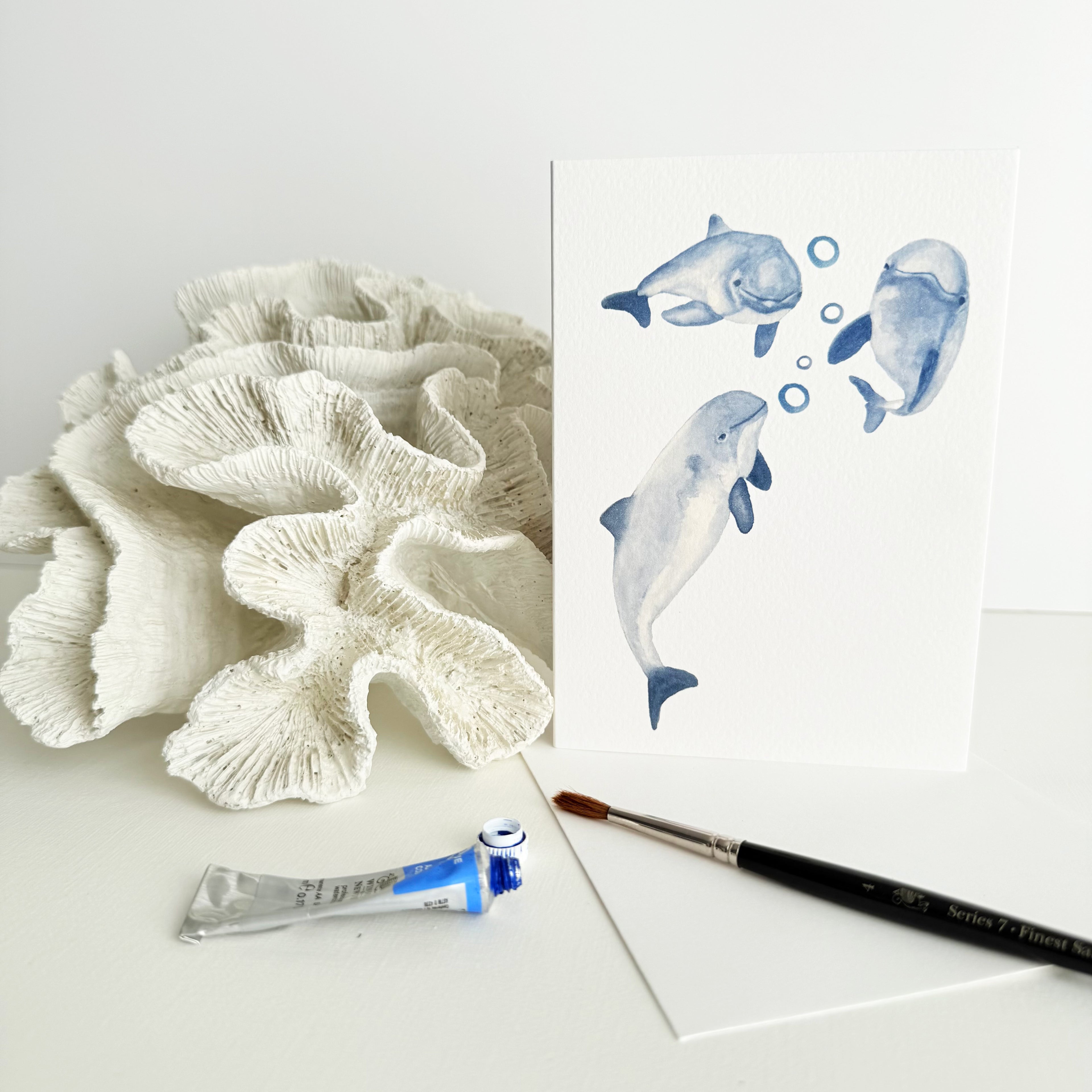 Artistic depiction of a greeting card with three porpoises blowing bubbles leaning against coral on a white surface with a paintbrush and tube of paint.