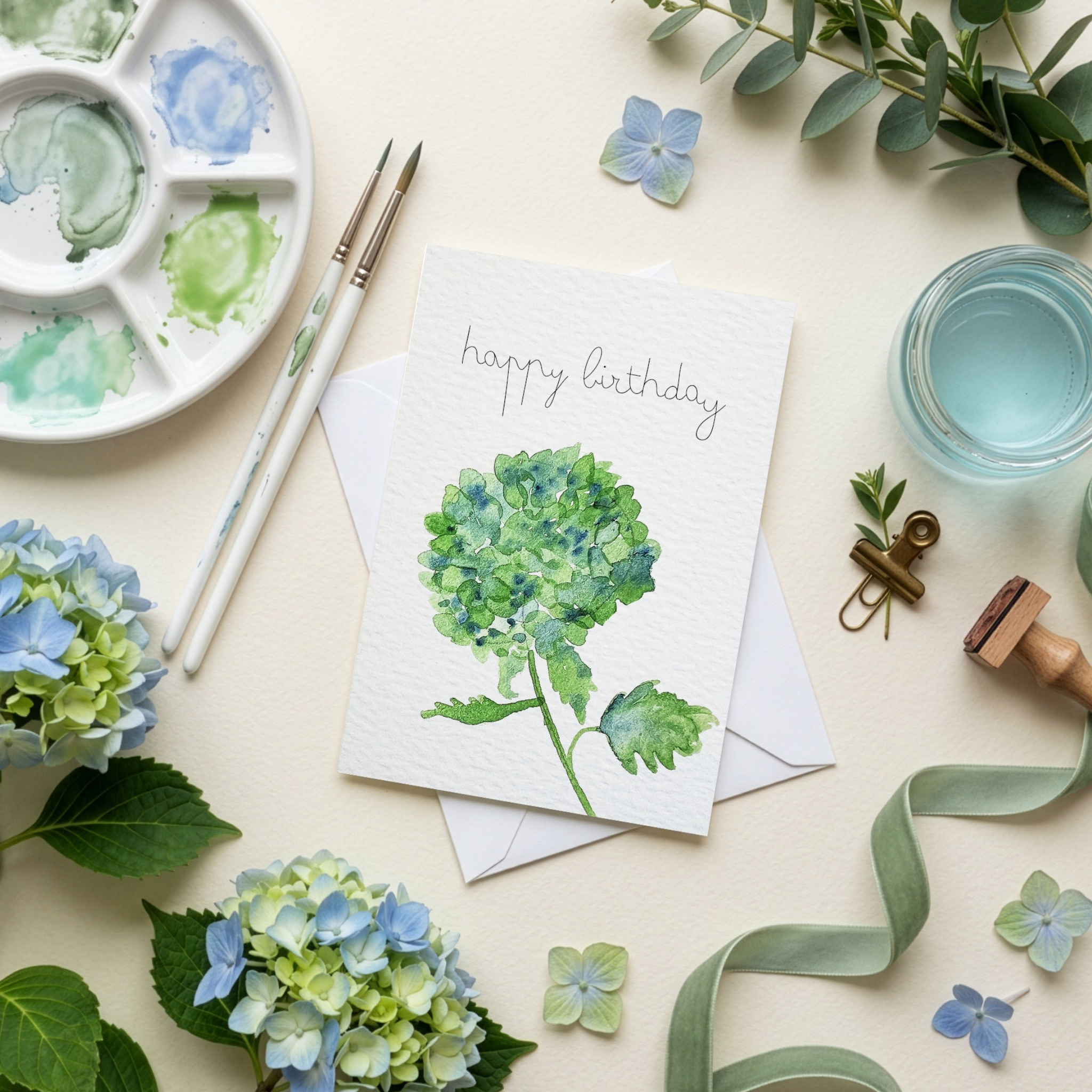 Greeting card with watercolor flowers, paint palette, and stationery items on a light background