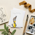Greeting card with a lemur watercolour illustration and 'happy birthday' text, surrounded by art supplies on a beige surface.