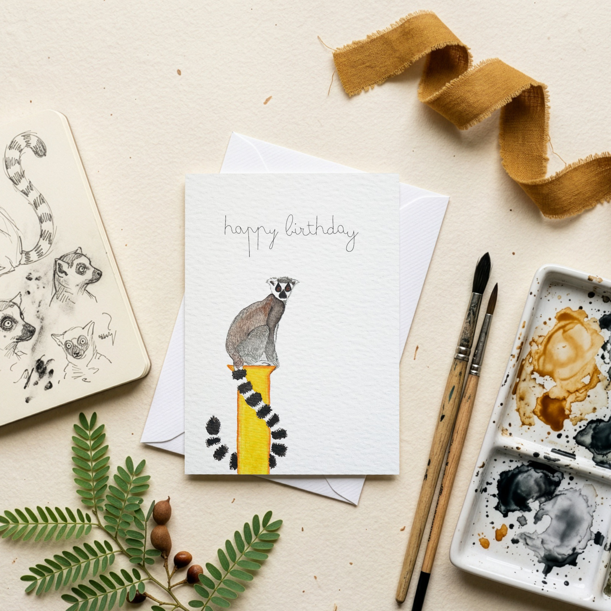 Greeting card with a lemur watercolour illustration and 'happy birthday' text, surrounded by art supplies on a beige surface.