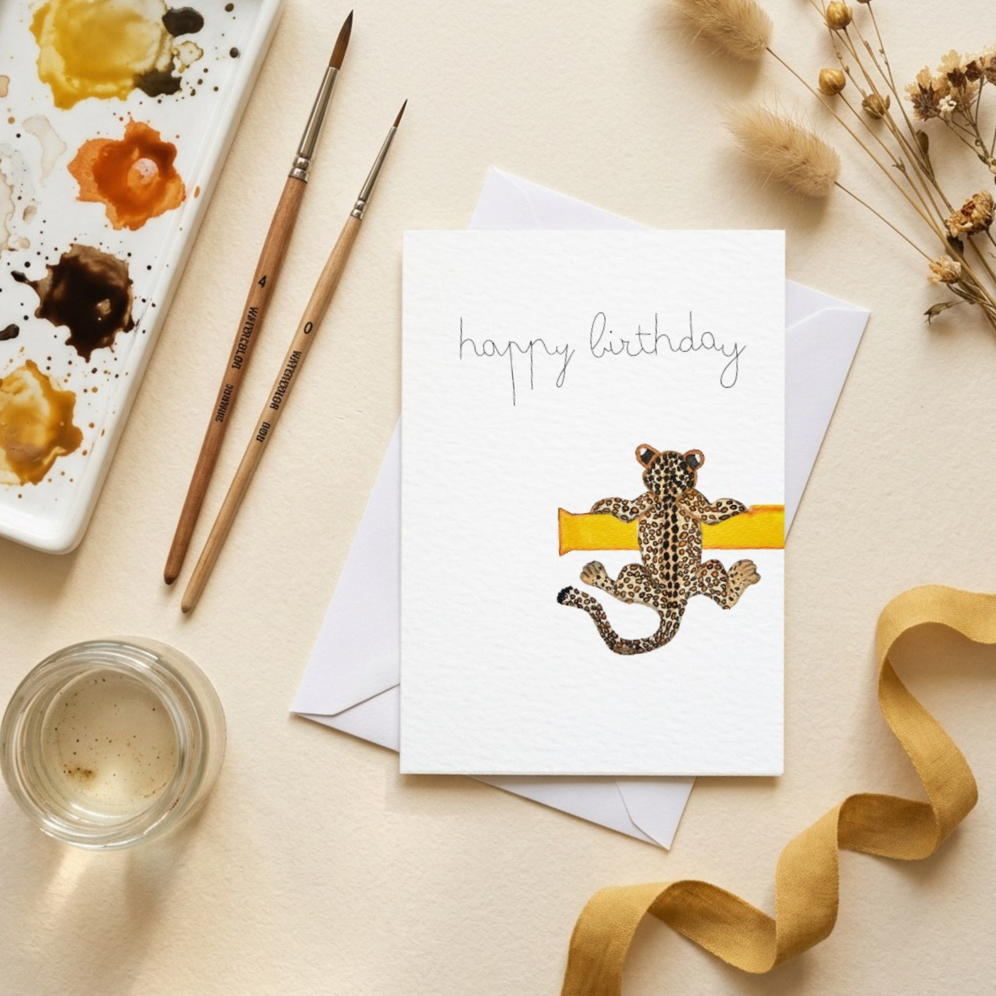 Birthday card with a watercolour illustration of a leopard cub hanging of a yellow ledge on a beige surface with art supplies