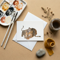 Artistic setup with watercolour palette, brushes, and a watercolour greeting card illustration showing a leopard mum with her cub on a beige surface.
