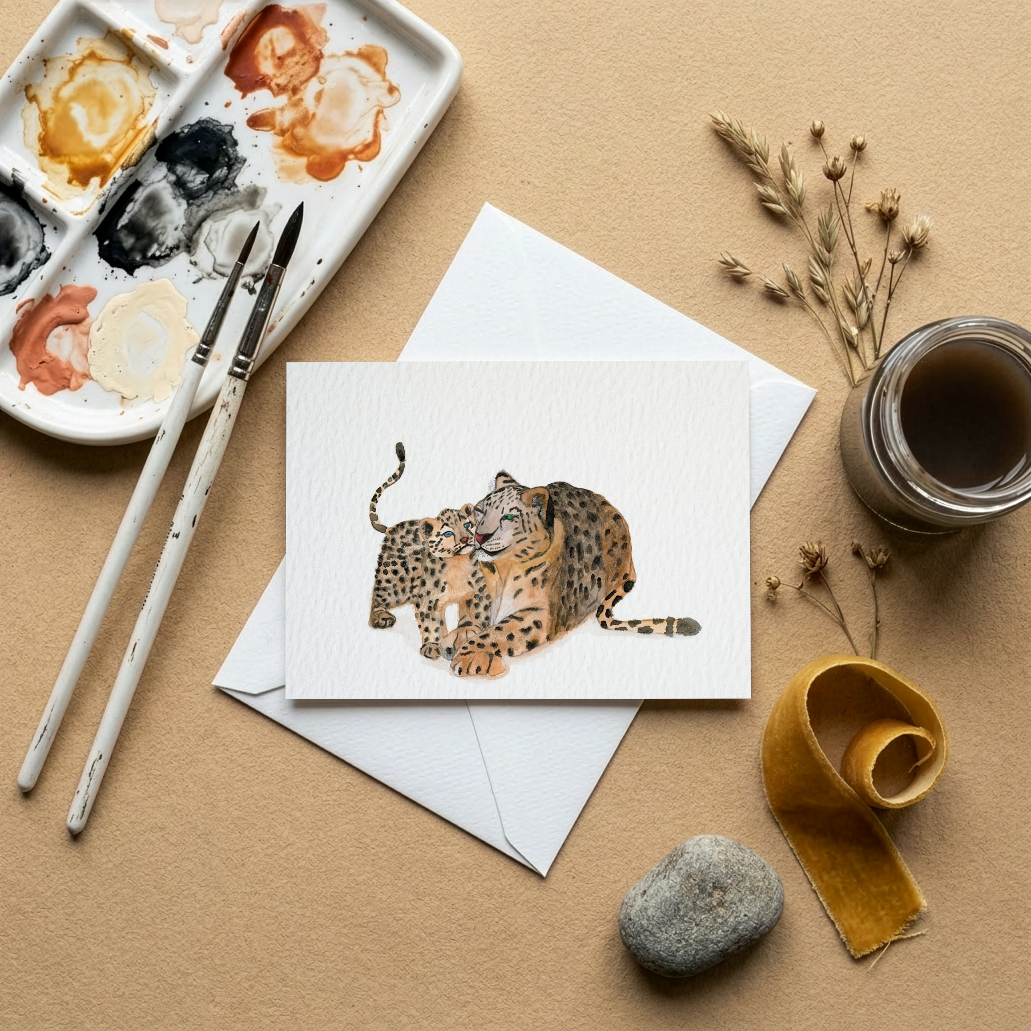 Artistic setup with watercolour palette, brushes, and a watercolour greeting card illustration showing a leopard mum with her cub on a beige surface.