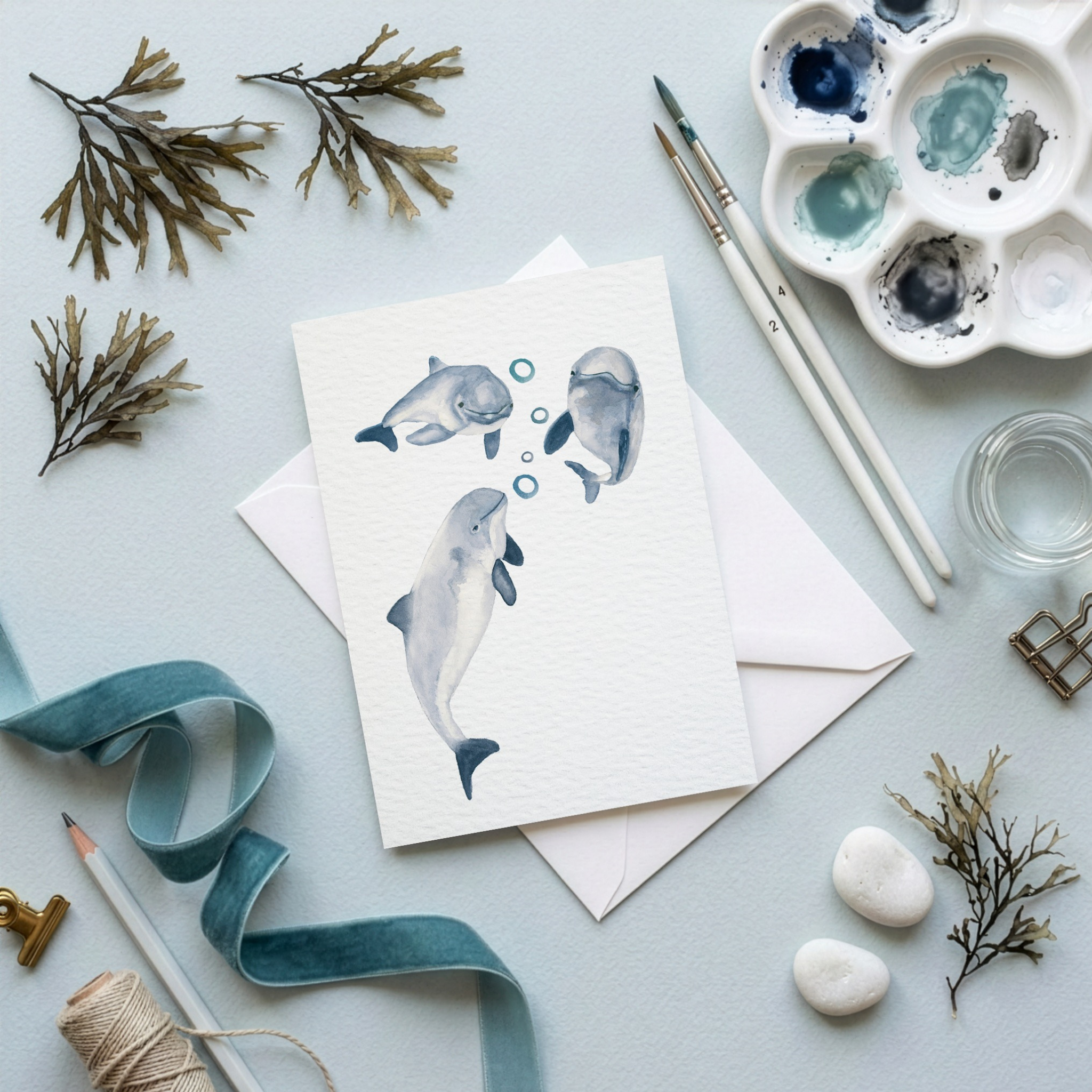 Greeting card with porpoise watercolour illustration, paint palette, brushes, and decorative elements on a light blue background