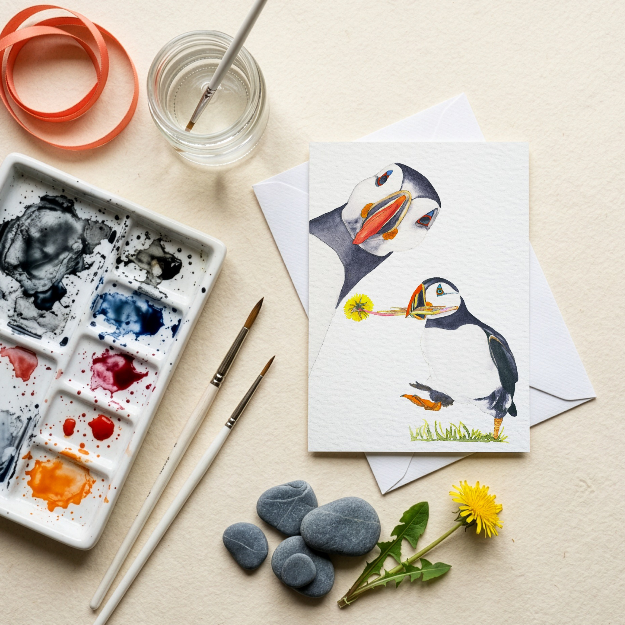 Art supplies including a watercolour palette, brushes, and a card with watercolour illustrated puffins on a beige surface.