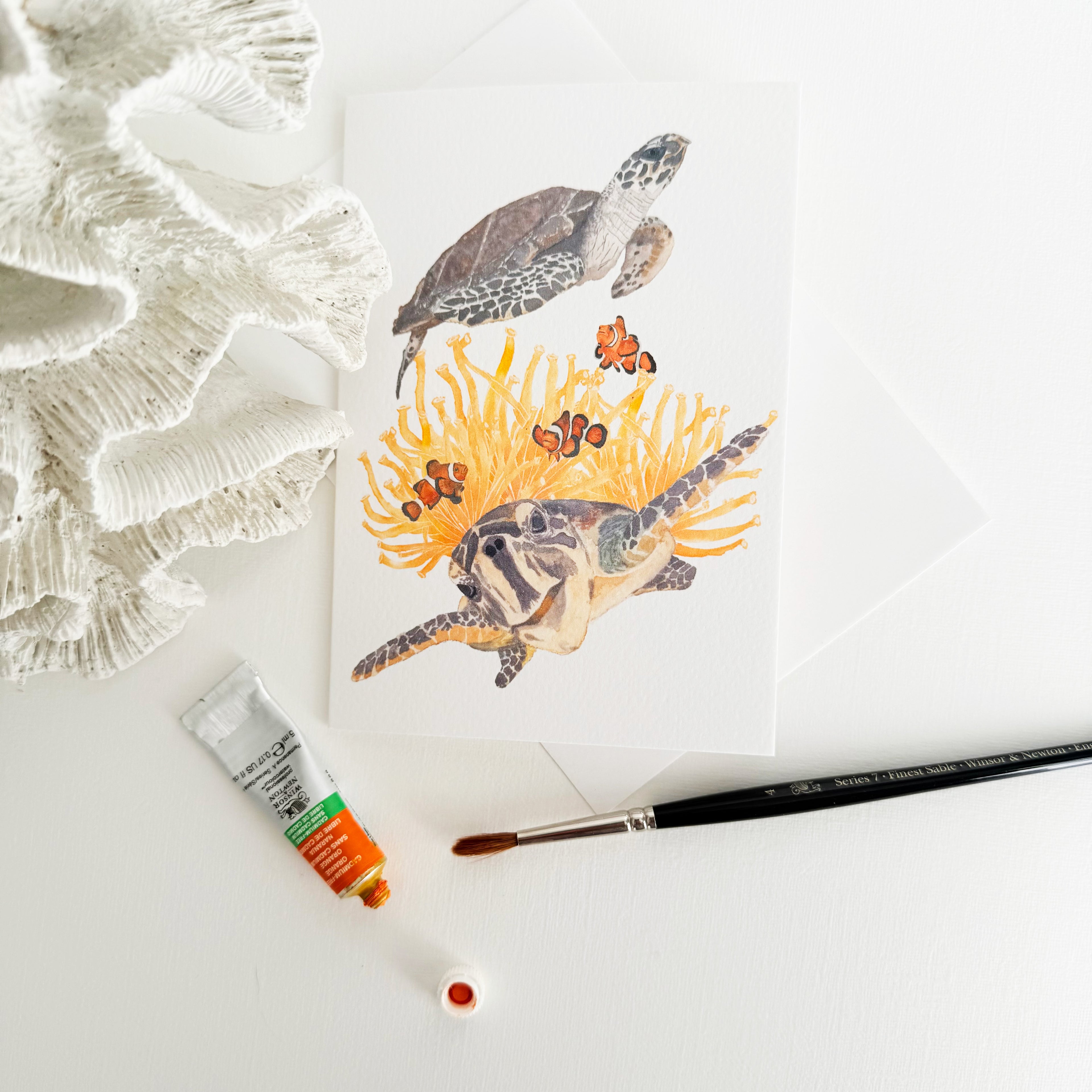Artistic depiction of a hand-illustrated greeting card of sea turtles leaning against coral with a paintbrush and paint tube on a white background
