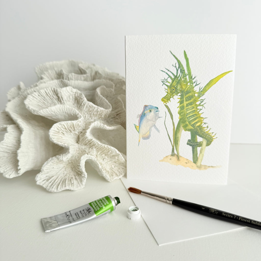 Artistic depiction of a watercolour hand-illustrated card of a green seahorse leaning against coral with a paintbrush and tube of paint on a white surface.