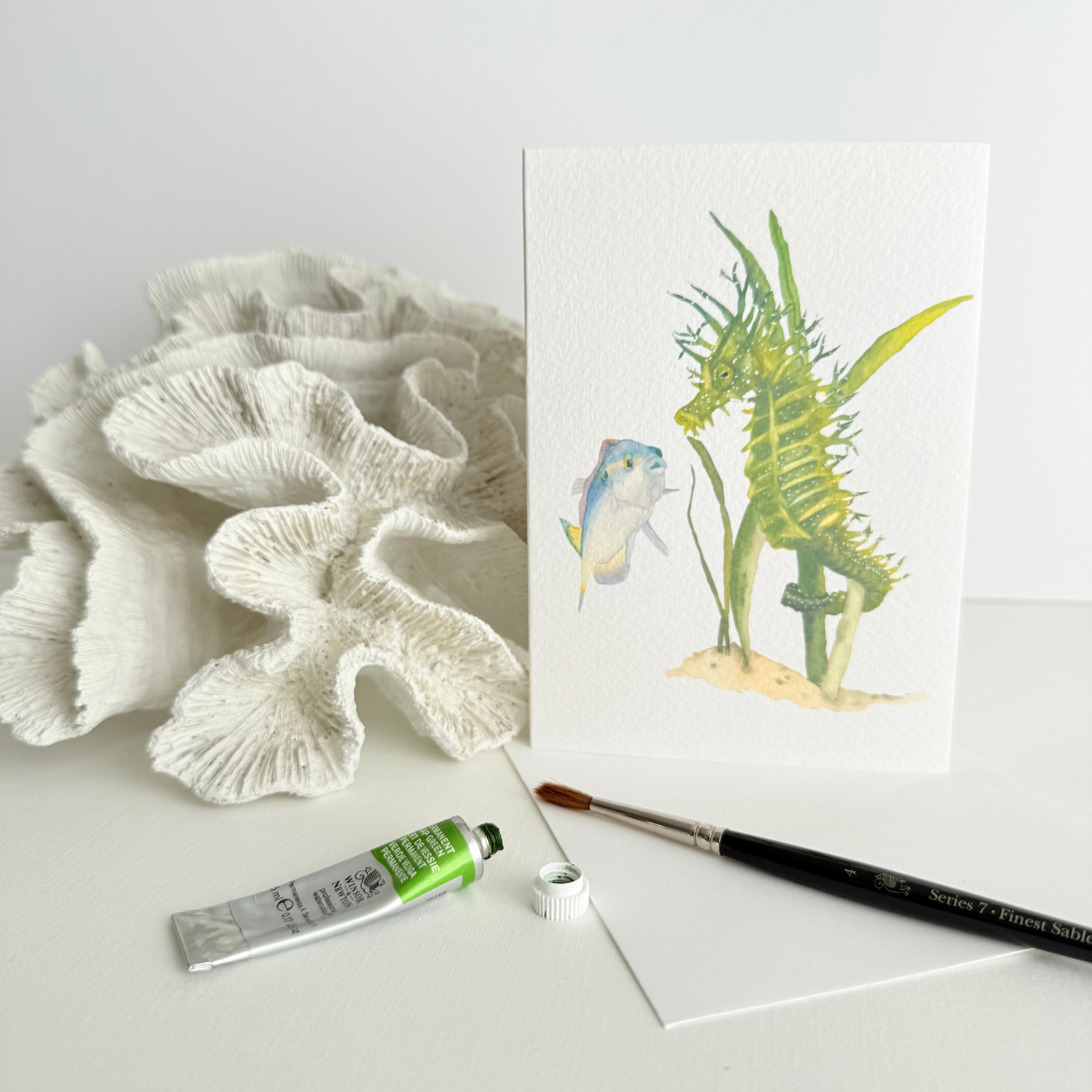 Artistic depiction of a watercolour hand-illustrated card of a green seahorse leaning against coral with a paintbrush and tube of paint on a white surface.