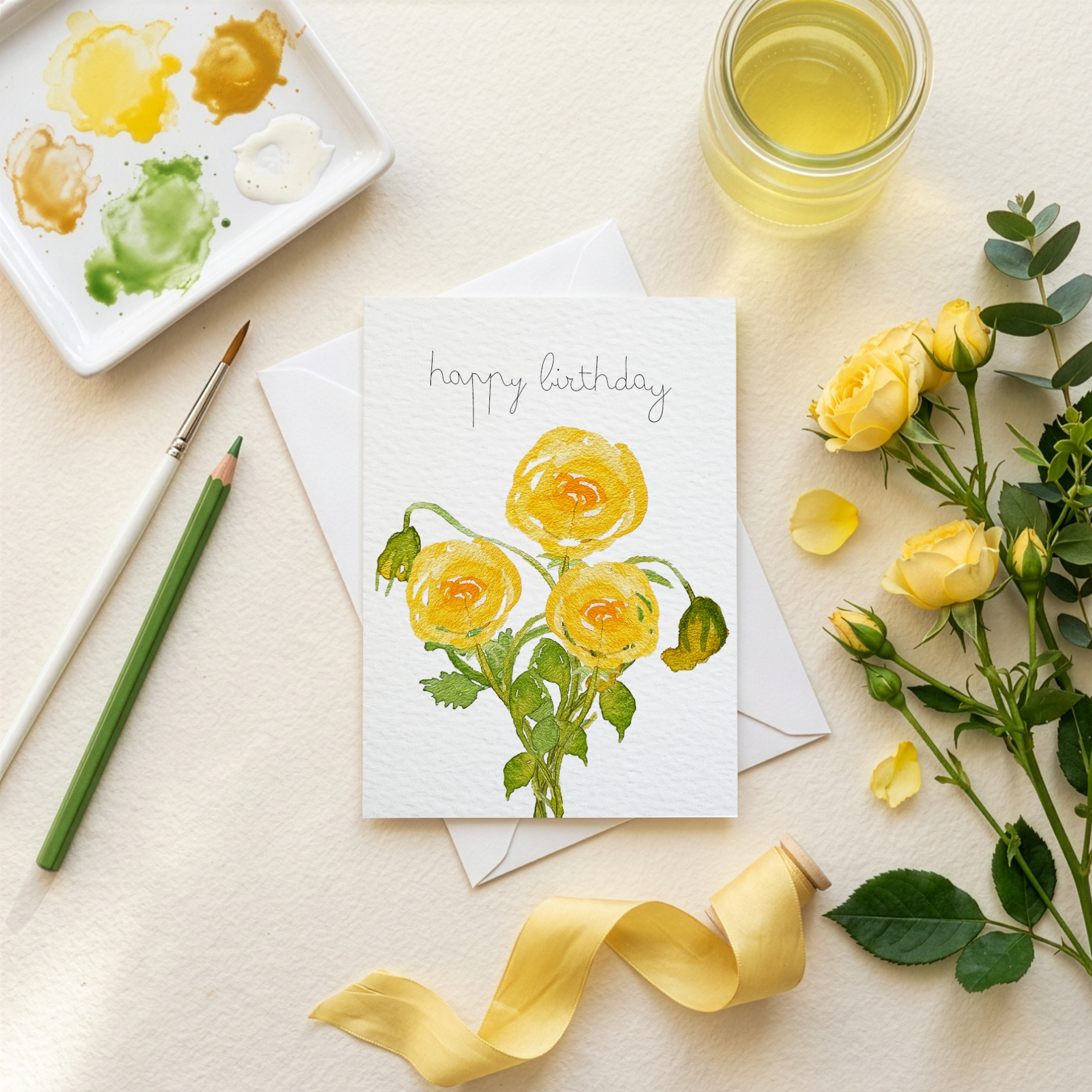 Birthday card with yellow watercolour illustrated roses, a paintbrush, and watercolor palette on a light surface.